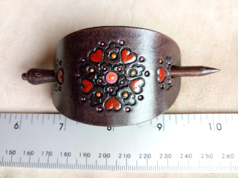 Może przedstawiać: Brązowa sk&oacute;rzana spinka do włos&oacute;w z ozdobnym wzorem w postaci pomarańczowych i r&oacute;żowych serc i kwiat&oacute;w. Spinka ma około 18 cm długości.