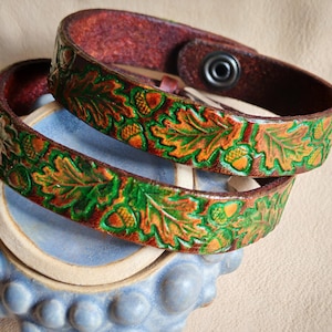 May include: Two brown leather bracelets with a carved oak leaf and acorn design. The leaves are painted green and orange, and the bracelets have a snap closure. The bracelets are stacked on a blue ceramic object.