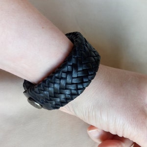K&ouml;nnte beinhalten: Ein schwarzes Lederarmband mit geflochtenem Design. Das Armband wird mit einem Metallverschluss geschlossen.