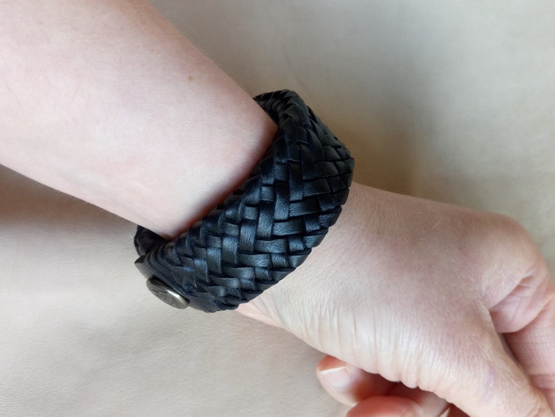 K&ouml;nnte beinhalten: Ein schwarzes Lederarmband mit geflochtenem Design. Das Armband wird mit einem Metallverschluss geschlossen.