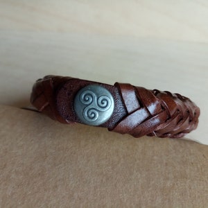 Puede incluir: Un brazalete de cuero marr&oacute;n con un dise&ntilde;o trenzado y un dije de metal plateado con un s&iacute;mbolo de triple espiral.