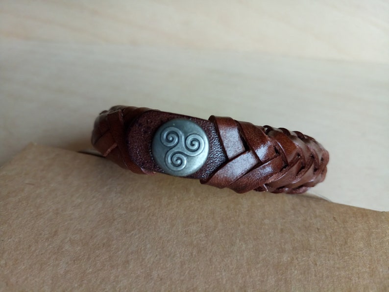 Puede incluir: Un brazalete de cuero marr&oacute;n con un dise&ntilde;o trenzado y un dije de metal plateado con un s&iacute;mbolo de triple espiral.