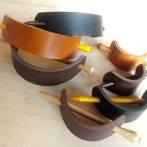 May include: A set of five leather hair clips with wooden sticks. The clips are in brown, black, and tan leather. The sticks are a light brown color.