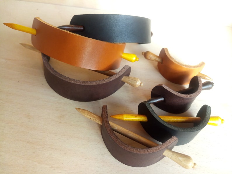 May include: A set of five leather hair clips with wooden sticks. The clips are in brown, black, and tan leather. The sticks are a light brown color.