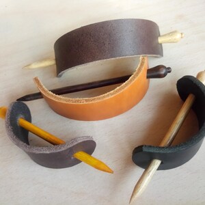 May include: Four leather hair clips with wooden dowels. The clips are in brown, tan, black, and gray. The dowels are in various shades of brown.