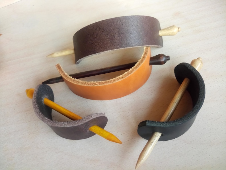 May include: Four leather hair clips with wooden dowels. The clips are in brown, tan, black, and gray. The dowels are in various shades of brown.