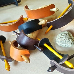 May include: Assortment of leather hair clips with wooden dowels. The clips are in various colors, including brown, black, and orange. The dowels are all yellow.