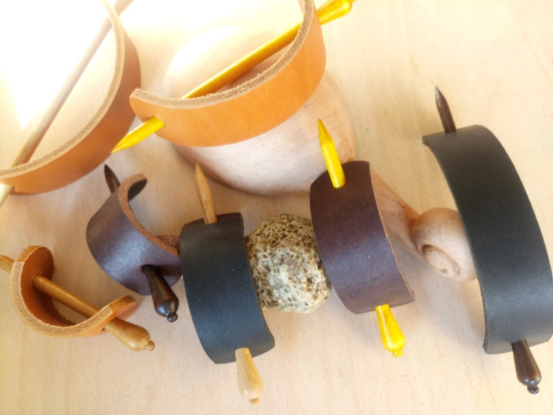 May include: Assortment of leather hair stick holders in brown, black, and dark brown with wooden hair sticks. Some sticks are yellow, brown, and dark brown.