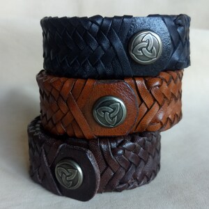 K&ouml;nnte beinhalten: Drei Lederarmb&auml;nder, die &uuml;bereinander gestapelt sind. Die Armb&auml;nder sind geflochten und haben vorne ein Metall-Keltenknotendesign. Das obere Armband ist schwarz, das mittlere Armband ist braun und das untere Armband ist dunkelbraun.