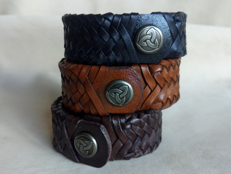K&ouml;nnte beinhalten: Drei Lederarmb&auml;nder, die &uuml;bereinander gestapelt sind. Die Armb&auml;nder sind geflochten und haben vorne ein Metall-Keltenknotendesign. Das obere Armband ist schwarz, das mittlere Armband ist braun und das untere Armband ist dunkelbraun.