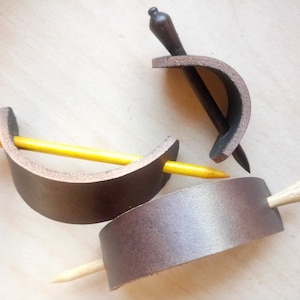 May include: Three leather hair clips with wooden hair sticks. The hair clips are brown and the hair sticks are yellow, brown, and natural wood.