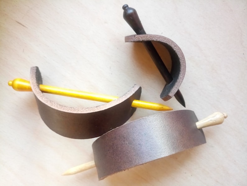 May include: Three leather hair clips with wooden hair sticks. The hair clips are brown and the hair sticks are yellow, brown, and natural wood.