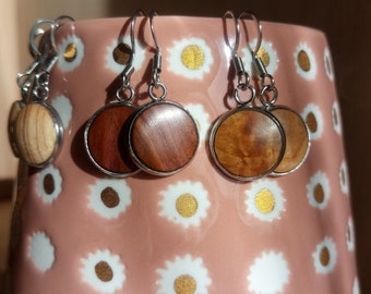 May include: Four pairs of earrings with wooden round discs in different shades of brown. The earrings are hanging from a pink surface with a white daisy pattern.