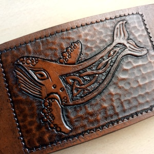 Może przedstawiać: Bransoletka ze skóry brązowej z wzorem celtyckiego węzła i wyrzeźbionym w skórze wielorybem.