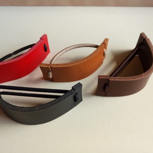 Op de afbeelding: Vier leren haarbandhouders in verschillende kleuren: rood, bruin, beige en zwart. Elke houder heeft een gebogen vorm en een enkel elastiekje.