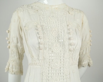 May include: A white cotton dress with intricate lace detailing and pom-pom accents. The dress has a high neckline and short sleeves.