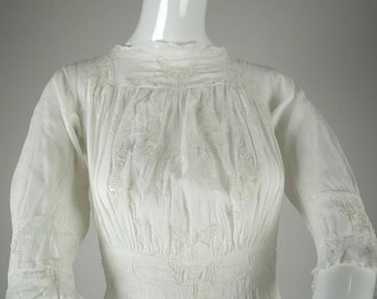 May include: White cotton dress with delicate embroidery throughout. The dress has a high neck and long sleeves.