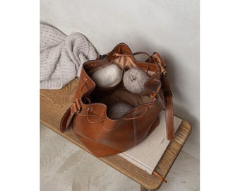 May include: A brown leather drawstring bag filled with yarn balls, resting on a wooden bench. The bag has a shoulder strap and is open, revealing the contents. A light-colored sweater is draped over the bench.