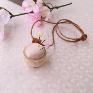 Puede incluir: Un cojín de alfileres de madera con una parte superior de tela rosa y un cordón de cuero marrón. El cojín de alfileres tiene forma de esfera y tiene tres alfileres con tapas de colores que sobresalen.