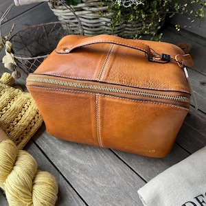 Può includere: Una borsa da viaggio in pelle marrone con cerniera e manico dorati. La borsa ha una forma rettangolare con cuciture e una piccola etichetta. La borsa è su una superficie di legno, lunga circa 25 cm.