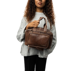 May include: A brown leather handbag with a top handle and a front pocket. The bag has a zipper closure and a rectangular shape. The leather has a textured appearance. The bag is held by a person wearing a gray sweater.