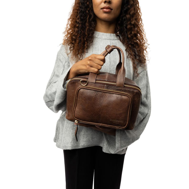 May include: A brown leather handbag with a top handle and a front pocket. The bag has a zipper closure and a rectangular shape. The leather has a textured appearance. The bag is held by a person wearing a gray sweater.