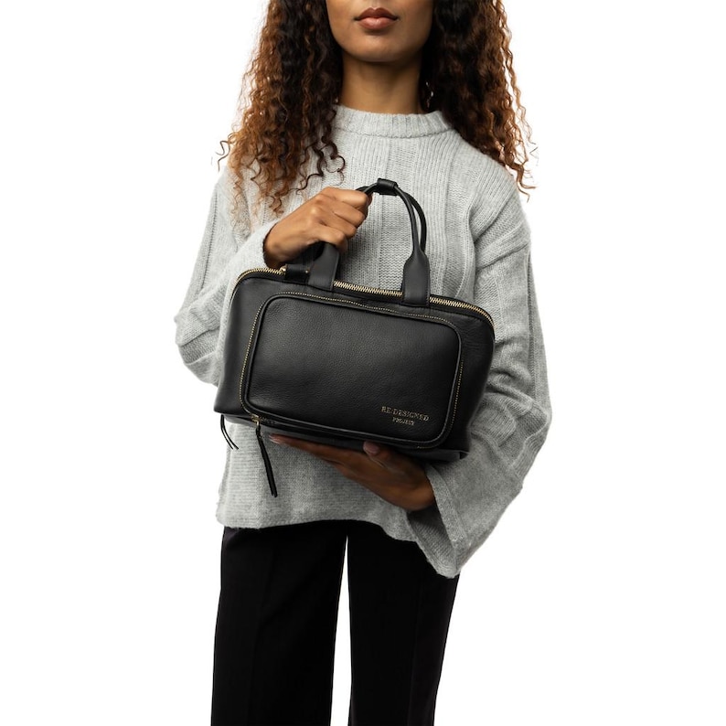 May include: A black leather handbag with a rectangular shape and a top handle. The bag features a zippered pocket on the front and gold-colored zippers. The text "BE REASONED PARIS" is embossed on the front of the bag.
