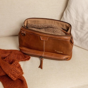 Può includere: Una borsa in pelle marrone con una cerniera aperta, che rivela un maglione marrone piegato. Una sciarpa lavorata a maglia color ruggine è vicina. La borsa ha un tirazip e il testo "RE DESIGNED" è visibile.