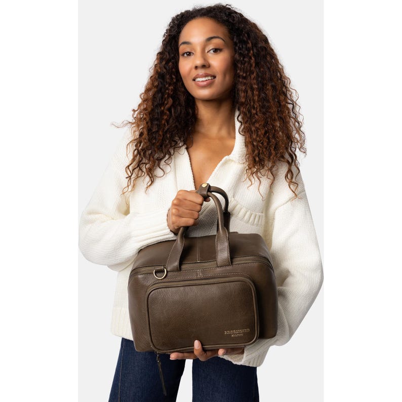 May include: A dark olive green leather handbag with a top handle and a front pocket. The bag has a rectangular shape and a zipper closure. The bag is held by a person wearing a white sweater and blue jeans.