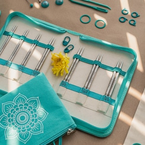 Peut inclure: Une trousse zippée bleu turquoise avec un intérieur blanc contient un jeu d'aiguilles à tricoter interchangeables. La trousse a un motif de mandala sur le devant. Les aiguilles sont disposées en rangées et étiquetées avec leurs tailles.