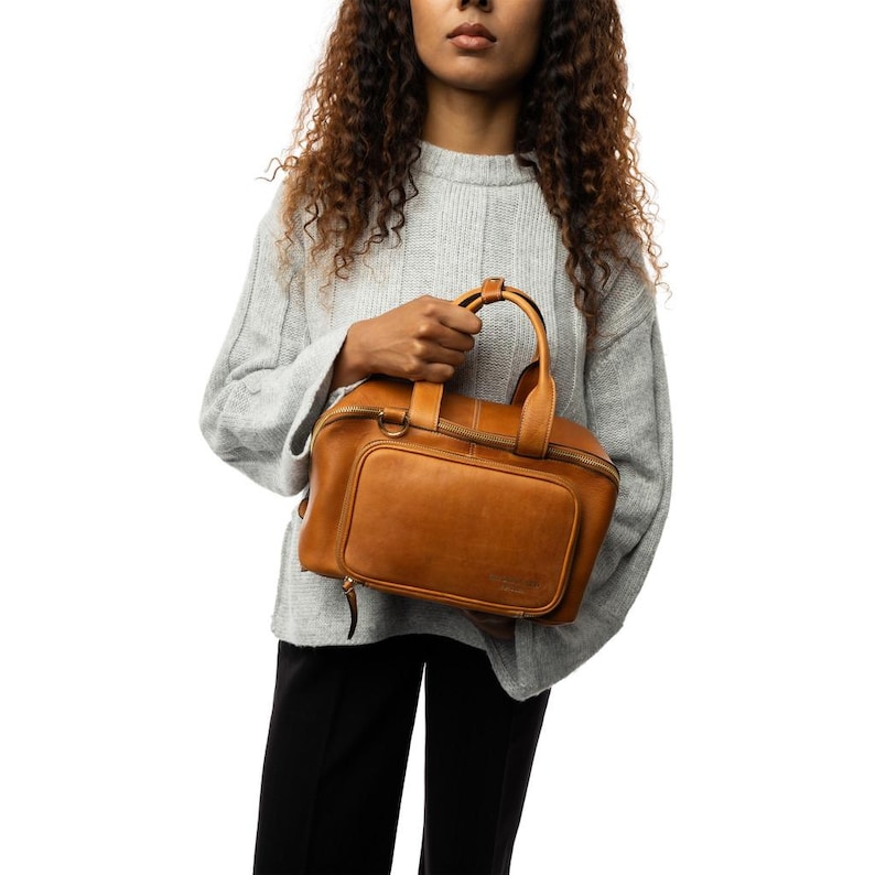 May include: A brown leather handbag with a top handle and a front pocket. The bag has a zipper closure and a rectangular shape. The bag is held by a person wearing a gray sweater and black pants.