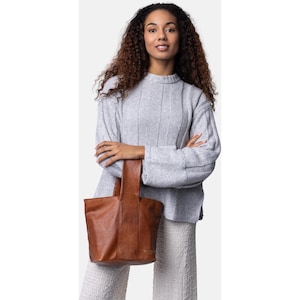 Può includere: Una donna tiene in mano una borsa tote in pelle marrone. La borsa ha un design strutturato con un unico manico. La donna indossa un maglione grigio chiaro a coste e pantaloni colorati chiari.
