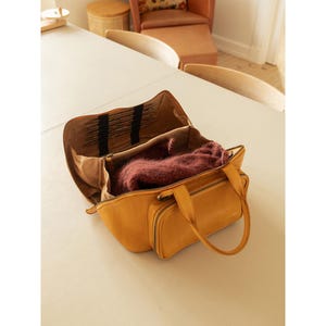 May include: Open mustard-yellow leather bag with a burgundy sweater inside. The bag has a top zipper, two front pockets with zippers, and a handle. The interior lining is tan with black accents.