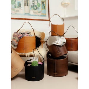Può includere: Una collezione di borse a secchiello cilindriche in pelle color cuoio, marrone scuro e nero. Ogni borsa presenta una tracolla singola, dettagli con cuciture verticali e una piccola etichetta rettangolare con la scritta RE:DESIGNED DENMARK.