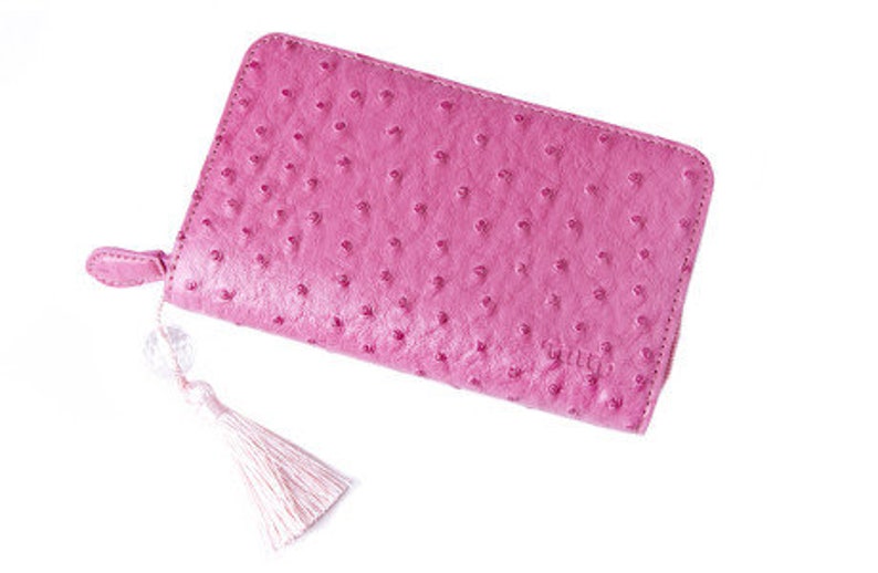 May include: A pink leather wallet with a zip closure and a tassel. The wallet has a textured surface with small, evenly spaced bumps. The tassel is white. The wallet is rectangular and appears to be new.