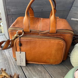 May include: A brown leather handbag with a top handle and a detachable shoulder strap. The bag has a front pocket with a zipper and a tag that reads "RE-DESIGNED PROJECT". The bag is sitting on a wooden surface.