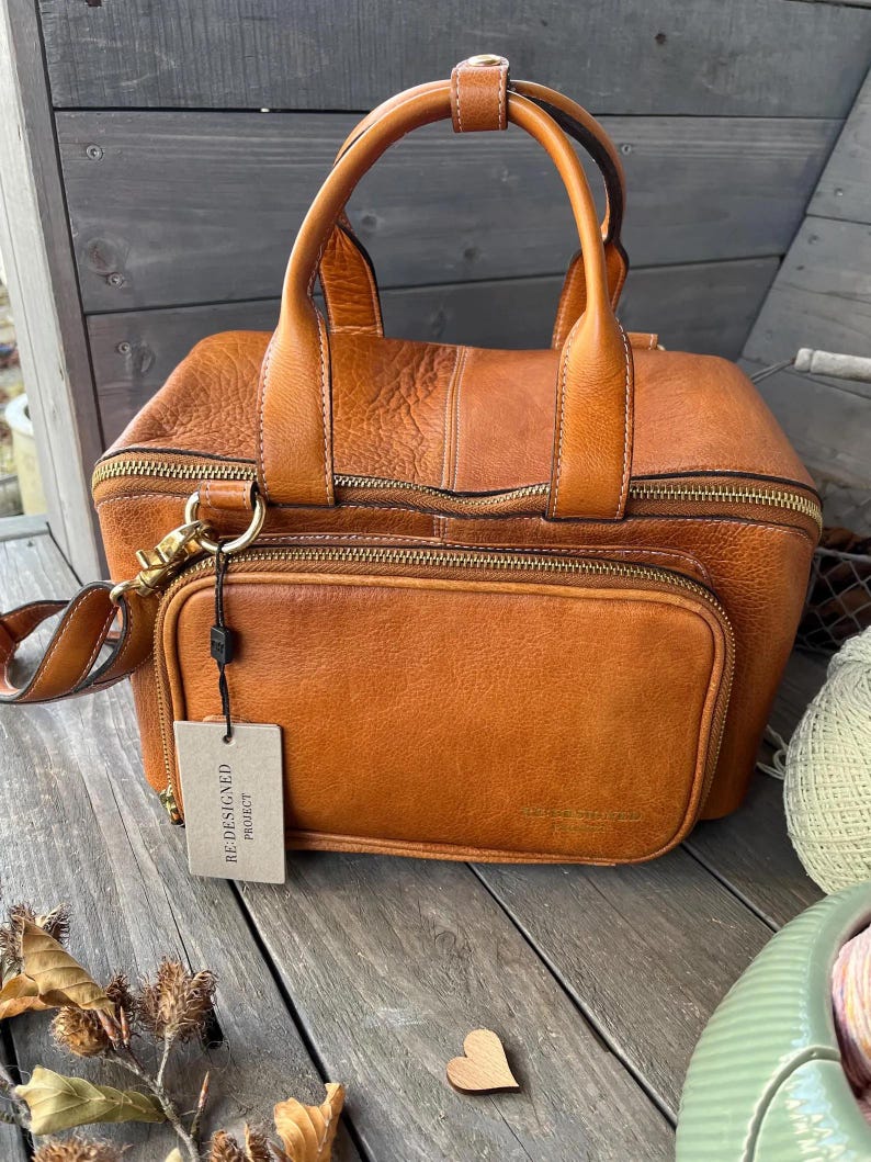 May include: A brown leather handbag with a top handle and a detachable shoulder strap. The bag has a front pocket with a zipper and a tag that reads "RE-DESIGNED PROJECT". The bag is sitting on a wooden surface.