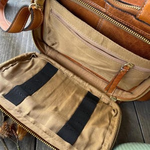 May include: Open leather briefcase with multiple compartments. The bag is a warm brown color with gold-colored zippers and hardware. The interior is tan with black elastic straps. A detachable shoulder strap is visible.