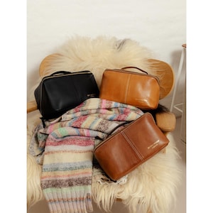 Può includere: Tre trousse da viaggio in pelle nera, marrone e beige con manici superiori e chiusure con cerniera. Una sciarpa a maglia a righe nei colori pastello poggia sotto le borse. Le borse sono su una sedia con una seduta soffice color crema.