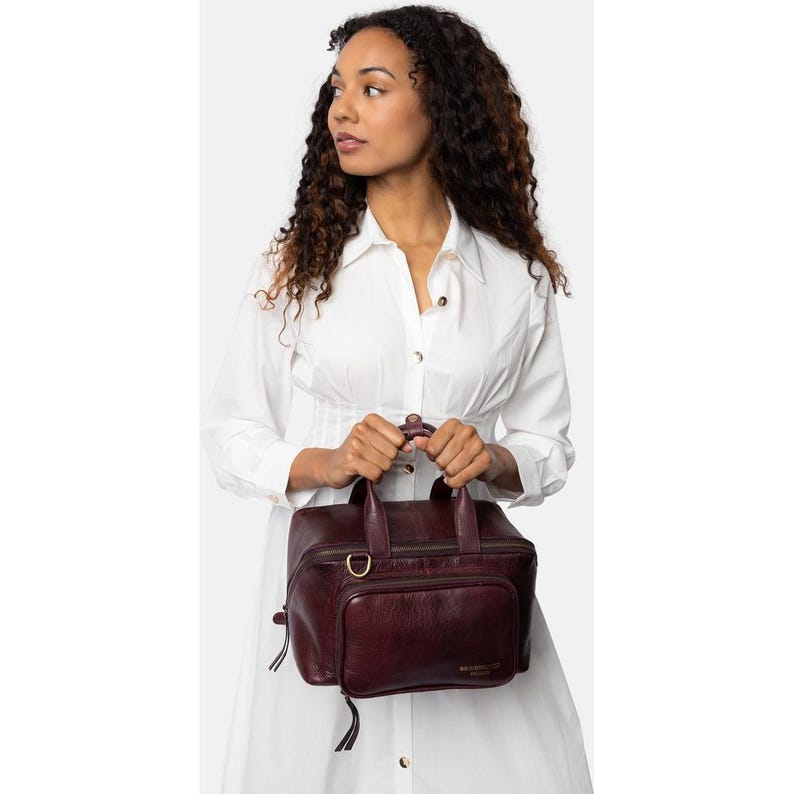 May include: A burgundy leather handbag with a top handle and a front pocket. The bag has a rectangular shape with a zipper closure and a gold-colored D-ring. The bag is held by a person wearing a white dress.