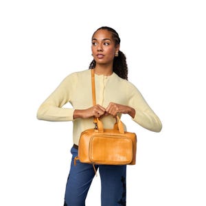 May include: A tan leather handbag with a shoulder strap and top handles. The bag has a rectangular shape with a front pocket and a zipper closure. The bag is held by a person wearing a yellow cardigan and blue jeans.
