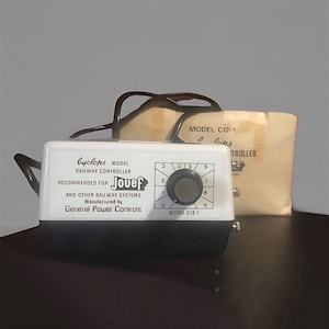 May include: A white and black Cyclops model railway controller with a dial and number indicators. The text on the controller reads "Jouef" and "Model C12-1". A tan paper package is behind the controller.