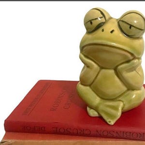 May include: A ceramic frog figurine with a contemplative expression, resting its chin on its hands. The frog is light green and sits atop a stack of red books with the title "Robinson Crusoe" visible.