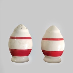 May include: A pair of ceramic salt and pepper shakers. Each shaker is egg-shaped, with a white base and a wide red stripe around the middle. The salt shaker has holes on top.
