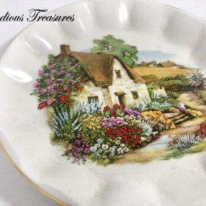 May include: A white, ruffled-edge decorative plate with a painted cottage scene. The cottage has a thatched roof and is surrounded by colorful flowers. The plate has a gold rim and the text "Melodious Treasures" is visible.