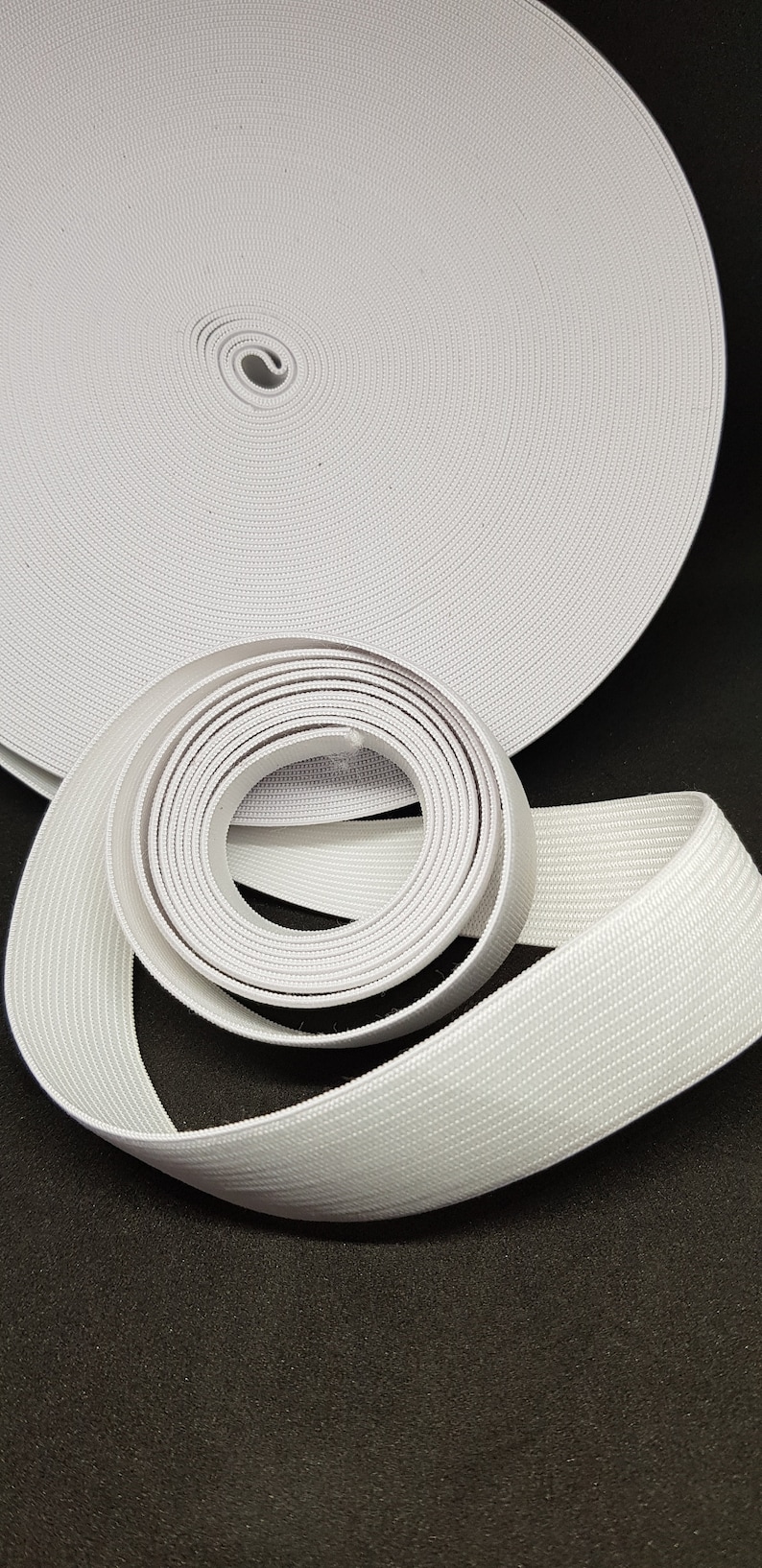 May include: White elastic band rolled up on a black surface. The elastic band is a flat, woven material with a textured surface.