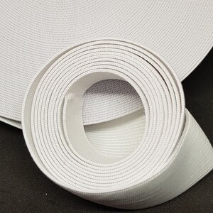 May include: A white elastic band rolled up on a black surface. The band is made of a woven material and has a textured surface.