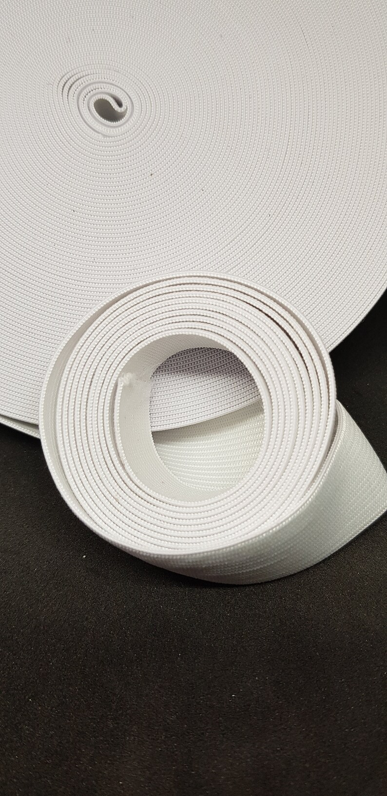 May include: A white elastic band rolled up on a black surface. The band is made of a woven material and has a textured surface.