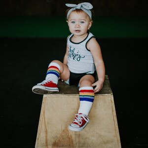 Op de afbeelding: Een jong kind draagt een grijs tanktop met de tekst "Little Warrior" erop gedrukt, zit op een houten kist. Het kind draagt regenboogkleurige, gestreepte kniekousen en rode sneakers.