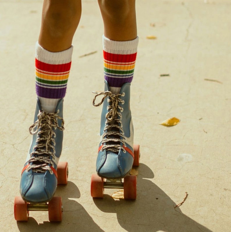Op de afbeelding: Een persoon draagt regenboogkleurige sokken en blauwe rollerskates met witte veters. De skates hebben rode en witte wielen.
